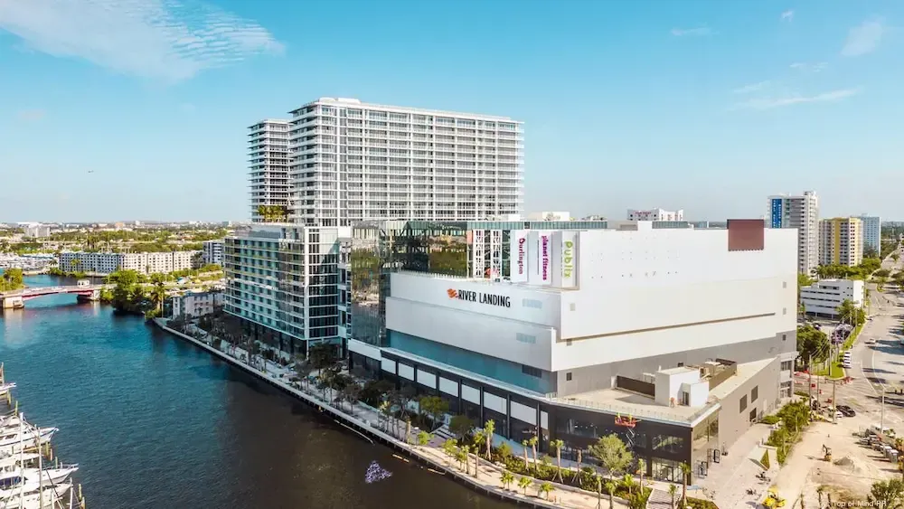 Modern waterfront shopping complex beside a canal, with a high-rise tower and blue sky.