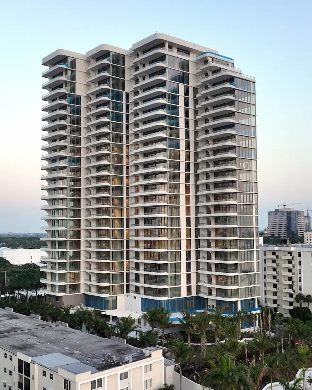 Tall modern high-rise apartment building with balconies, surrounded by smaller buildings and palm trees at dusk