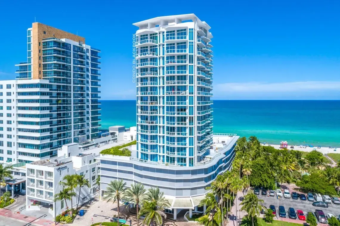 Modern beachfront high-rise condos with blue-tinted glass overlooking a turquoise ocean and palm-lined street