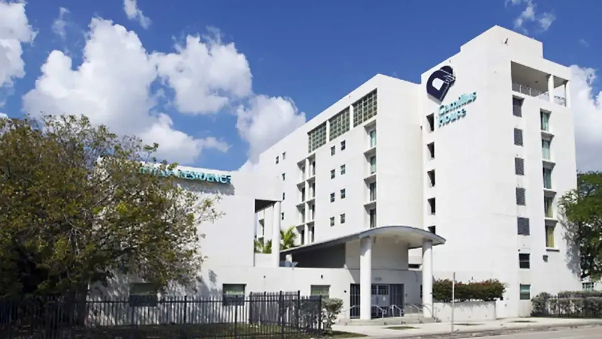 White multi-story building with a blue logo, trees in front, and a cloudy sky above
