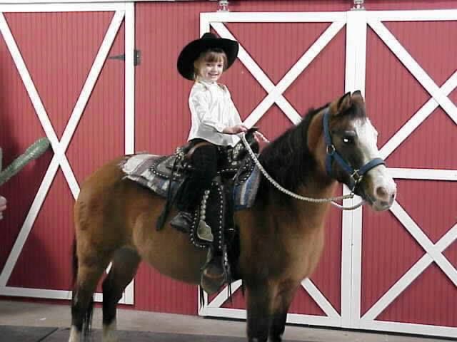 A smiling child in a cowboy hat and white shirt riding a brown pony in front of red barn doors with white trim.