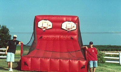 Two people stand beside a large, red, inflatable dual basketball hoop game set up in a field on a sunny day.