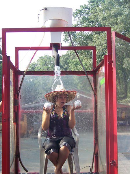 A person sits in a chair inside a red booth, wearing a straw hat while water pours down from a toilet tank above.