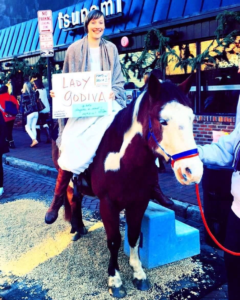 A smiling person sits on a brown-and-white horse in front of a building, holding a sign that reads 