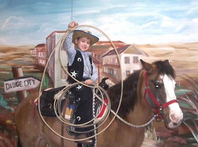 A person in a cowboy outfit holds a lasso while sitting on a horse in front of a painted backdrop of a western town.