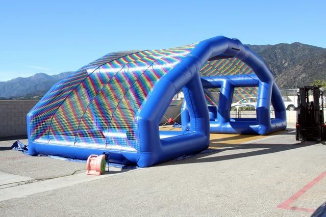 A large blue inflatable structure with a multicolored mesh top and open sides stands on a paved outdoor lot.