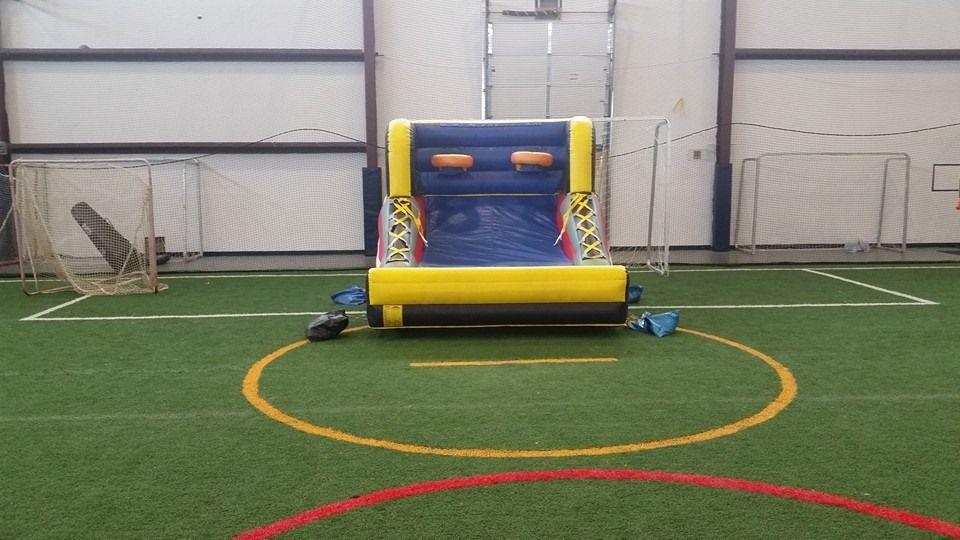 A yellow and blue inflatable basketball game sits on a green indoor sports field with soccer nets in the background.