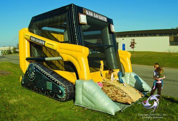 A yellow and black inflatable skid-steer bouncer on a grassy lawn with two people standing nearby.