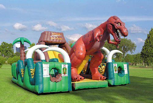 A red T-Rex inflatable obstacle course and slide set in a grassy park under a blue, cloudy sky.