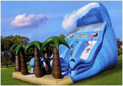 Inflatable blue wave slide with palm tree decorations on a grassy field under a bright sky.