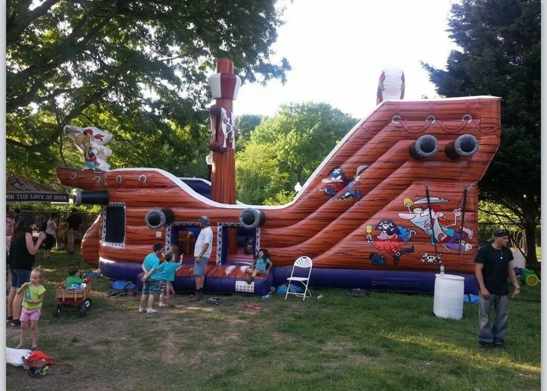 A large orange inflatable pirate ship in a park with several people standing nearby on the grass.