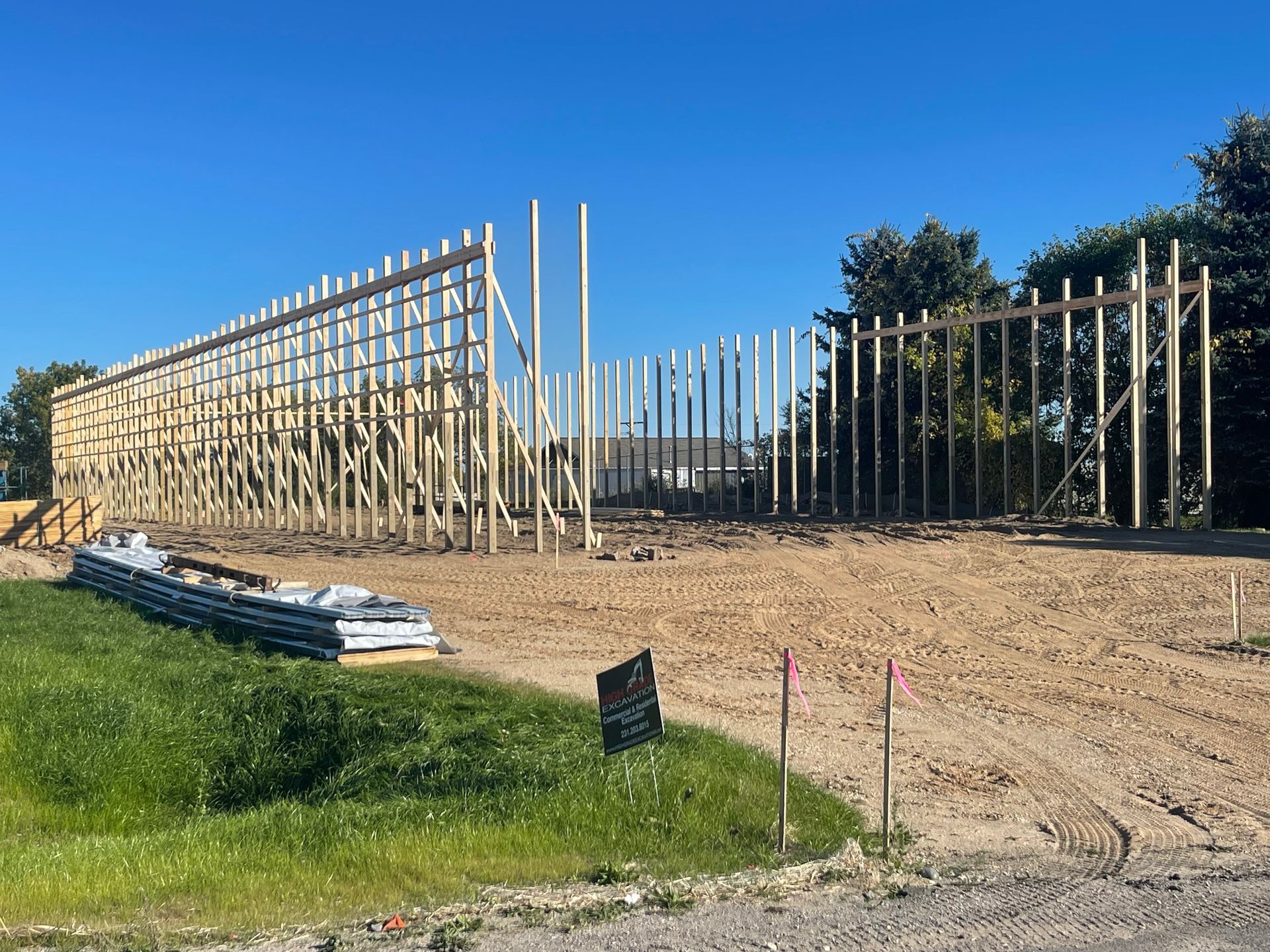 Wooden frame of a building under construction on a dirt lot, with a stack of sheet metal and some greenery.