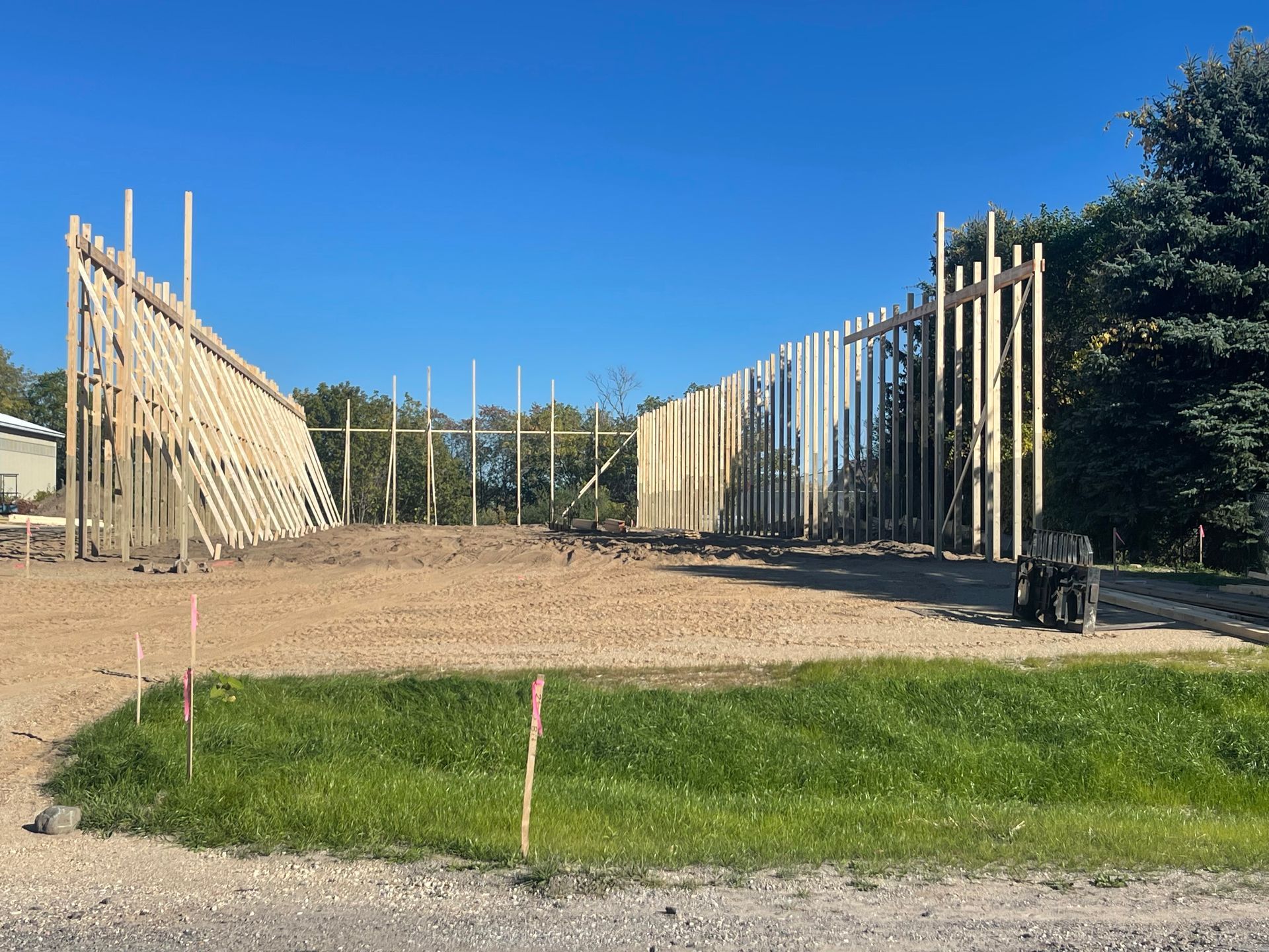 Framing of a new building under construction, wooden beams set against a blue sky, on a dirt and gravel lot.