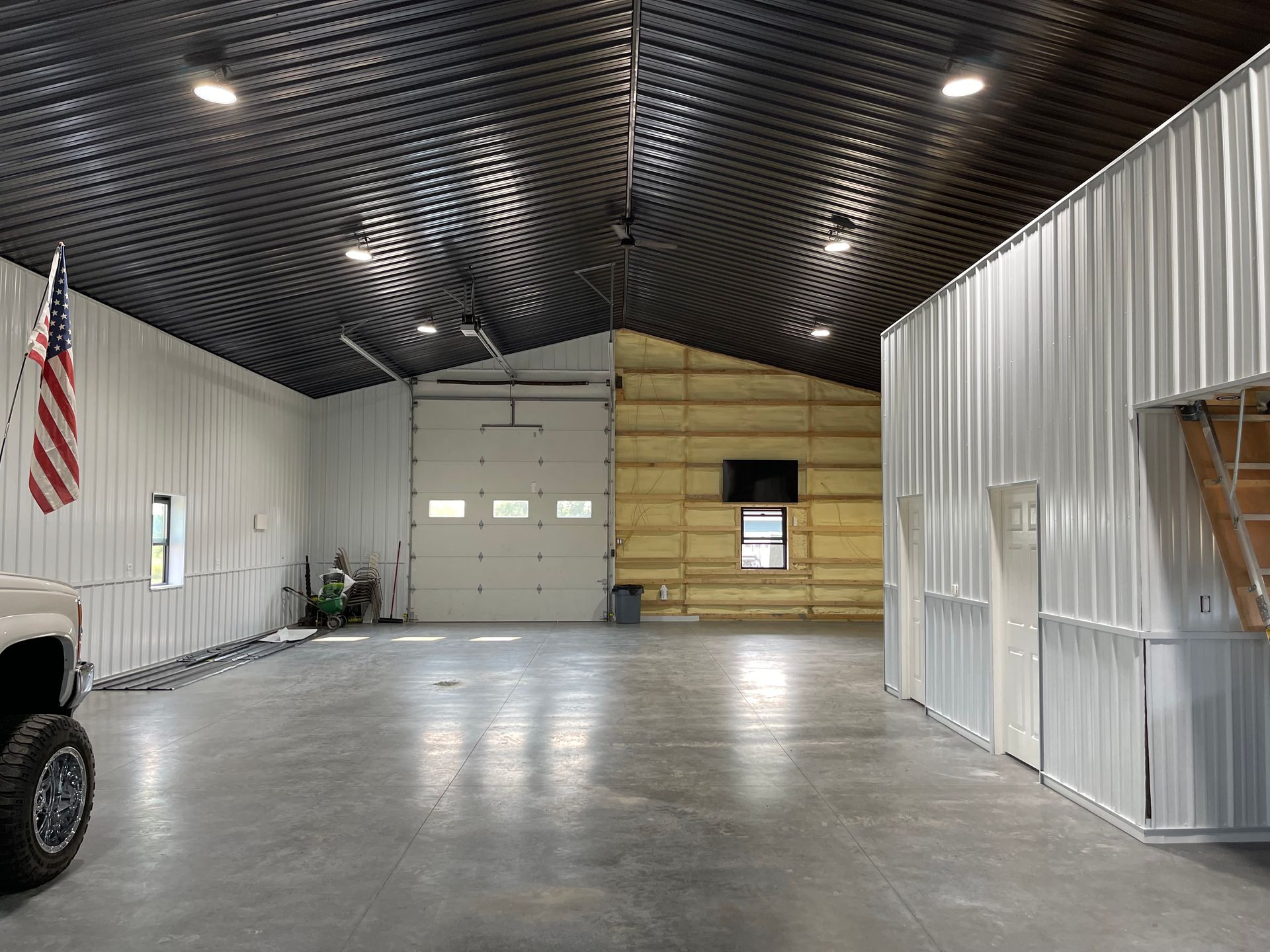 Interior of a large metal building. Garage door, concrete floor, white metal walls, black corrugated ceiling, American flag.