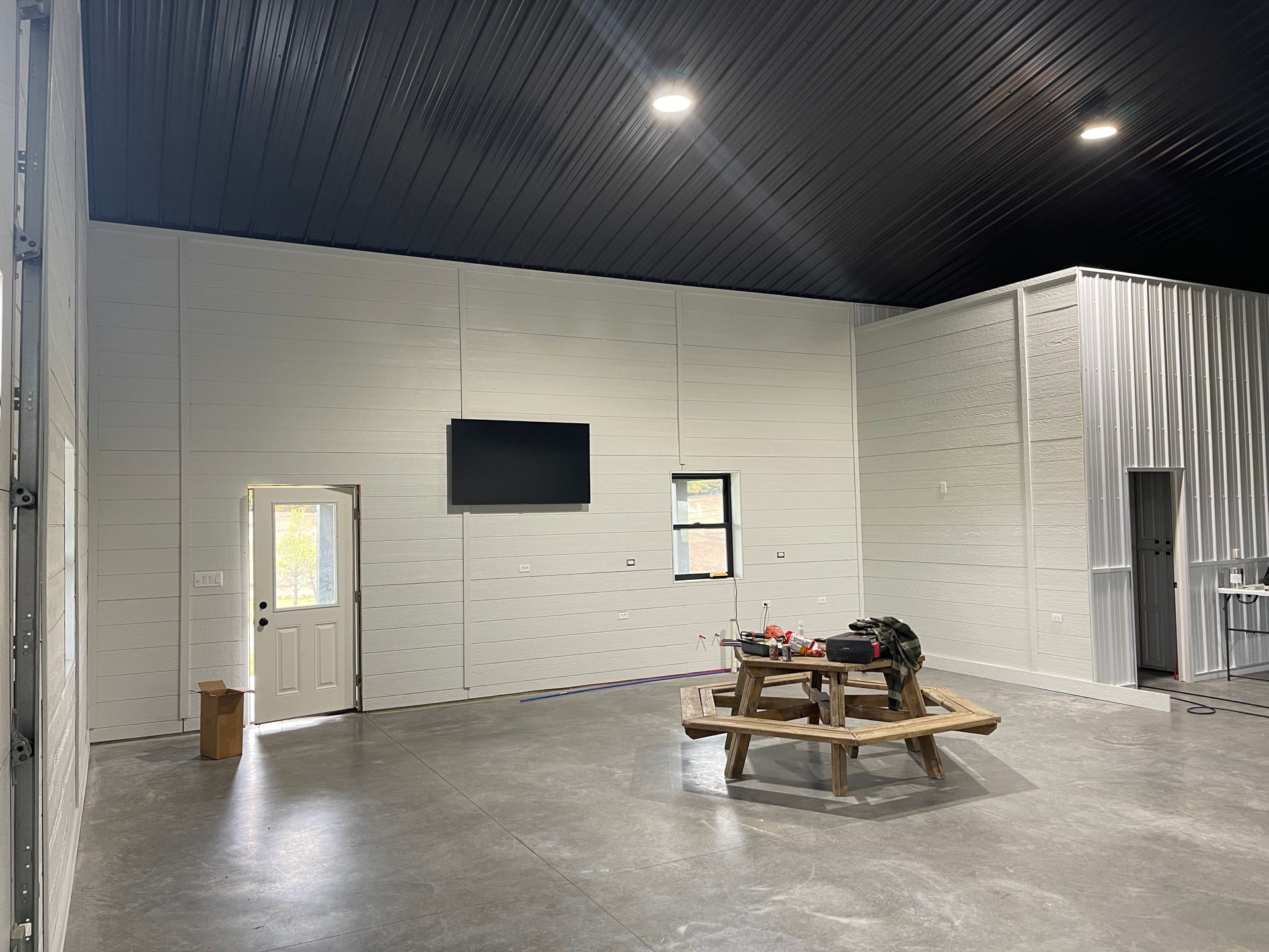 Spacious interior with white panel walls, black ceiling, concrete floor, wooden picnic table, TV, and door.