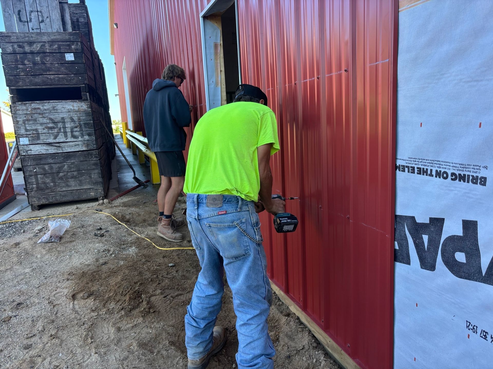 Two workers installing red metal siding on a building. One uses a tool, the other watches. Outdoors.
