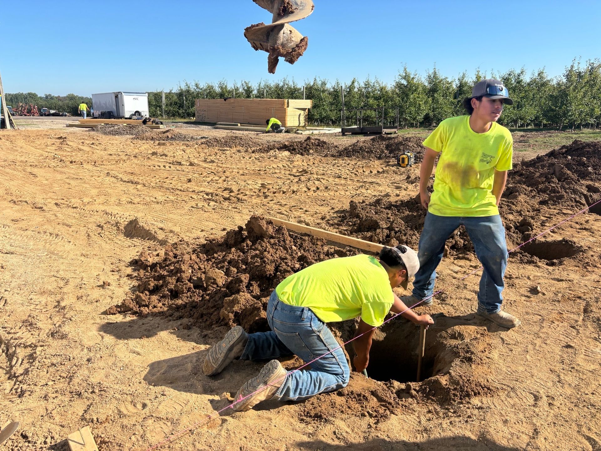 Two workers in neon shirts dig a hole, one looking in, one standing nearby, with a drilling device overhead.