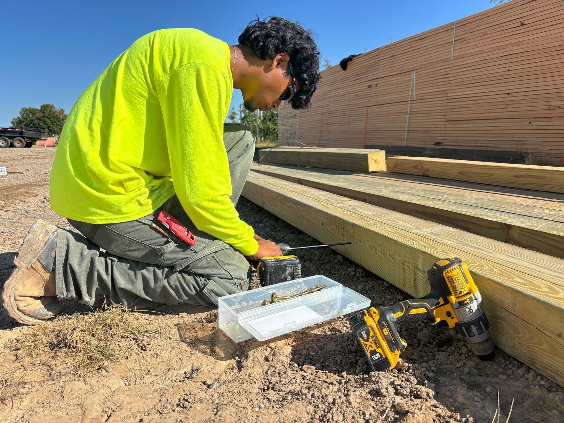 Construction worker kneeling, using a drill on lumber outside. Wearing neon yellow shirt.