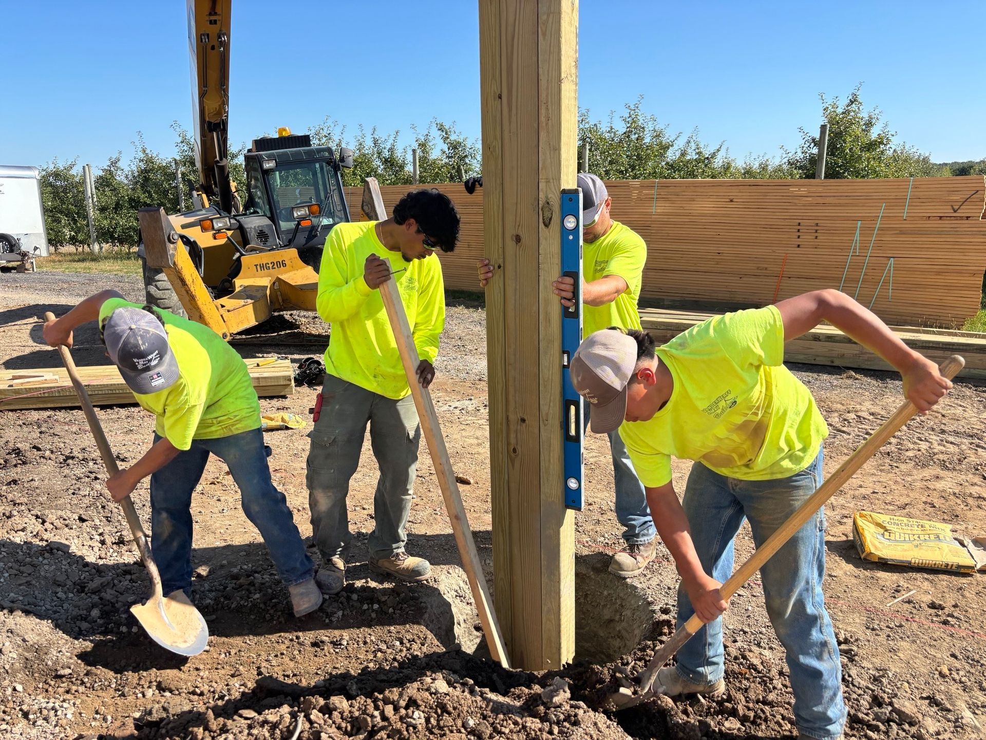 Four construction workers in neon shirts digging around a wooden pole outdoors; a yellow excavator in the background.
