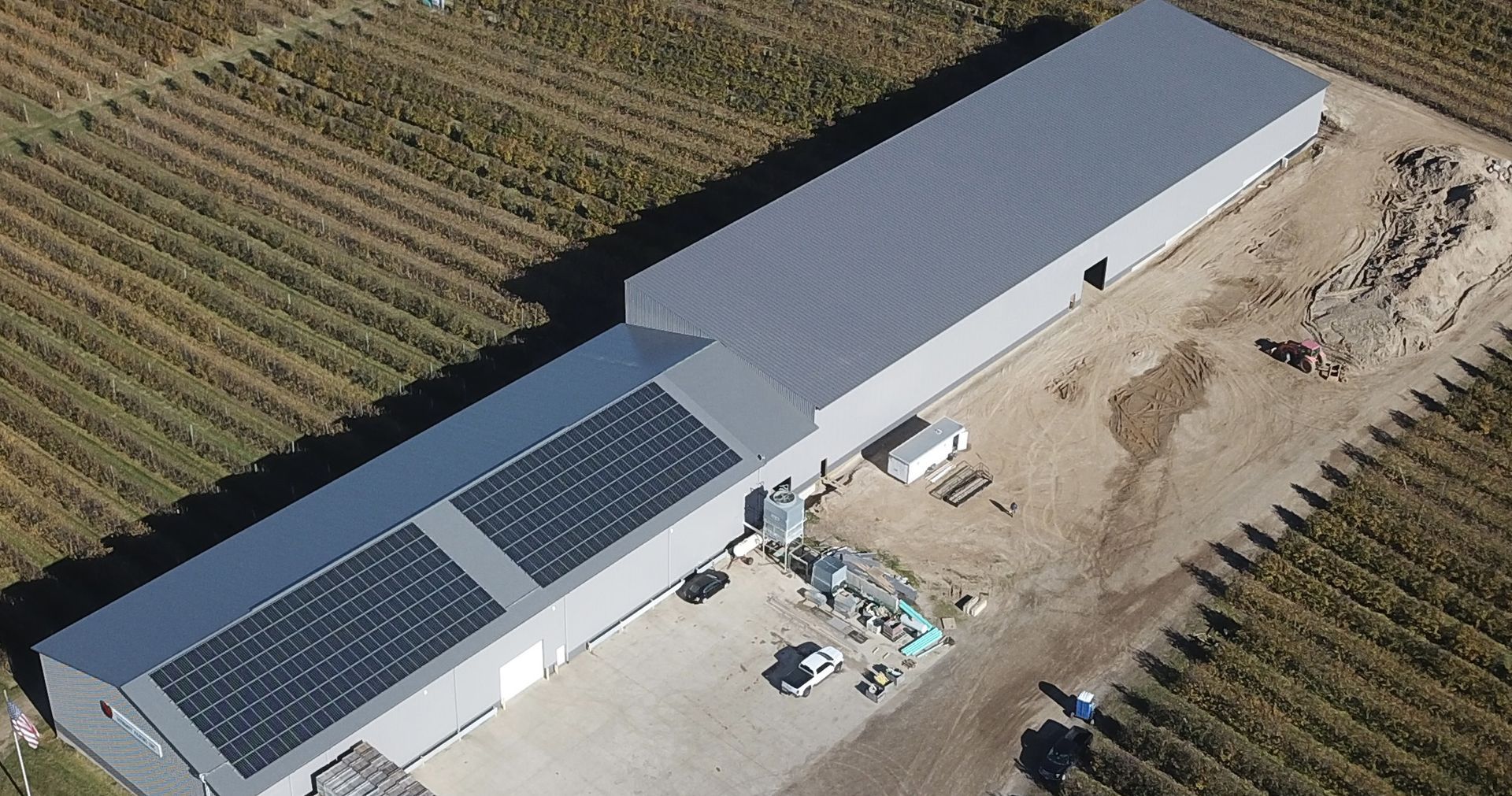 Aerial view of a long, gray building with solar panels, adjacent to a vineyard.