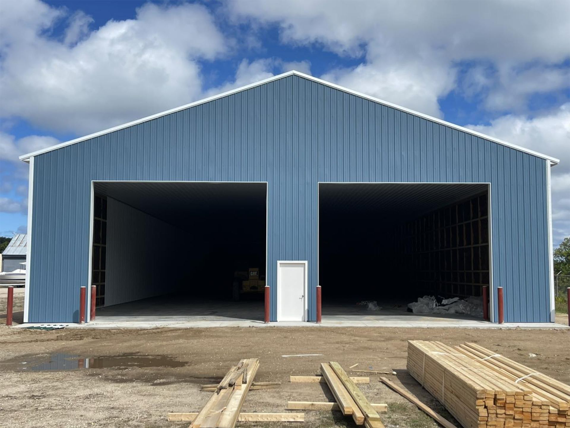 Blue metal building with two open bays and a white door; lumber in foreground, cloudy sky.