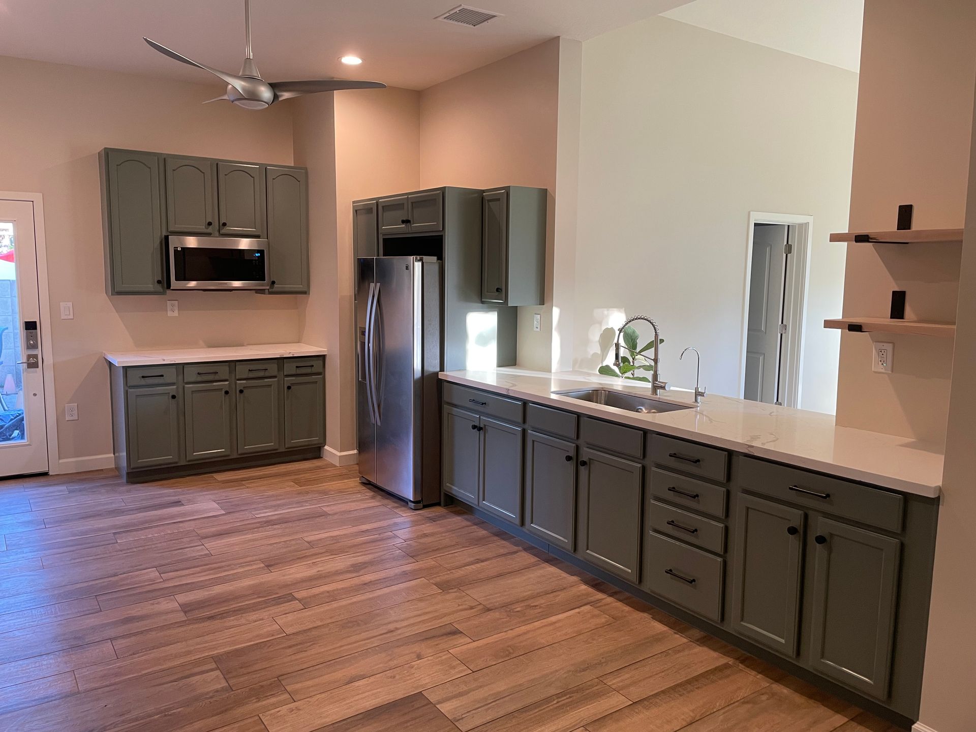 A kitchen with gray cabinets , a stainless steel refrigerator , a sink , and a ceiling fan.