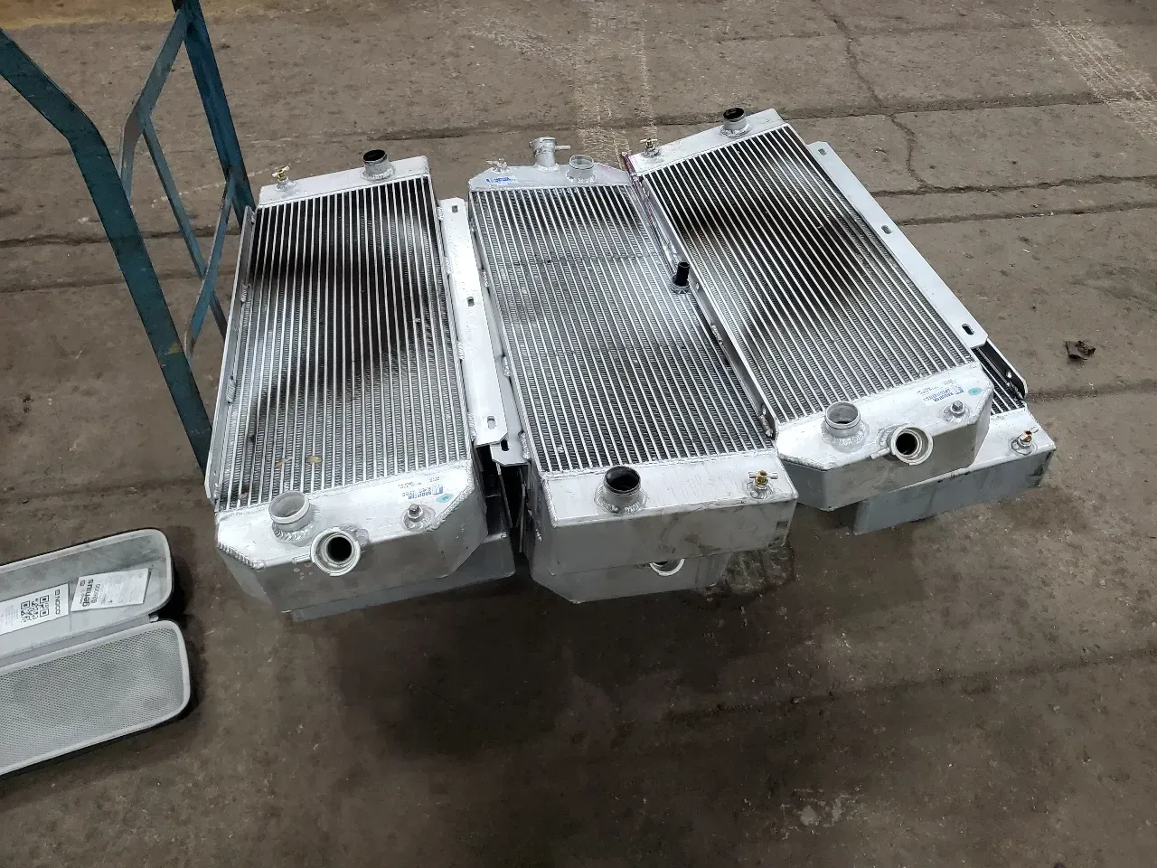 Three aluminum radiators, possibly for vehicles, sit on a gray concrete floor, leaning against a metal cart.