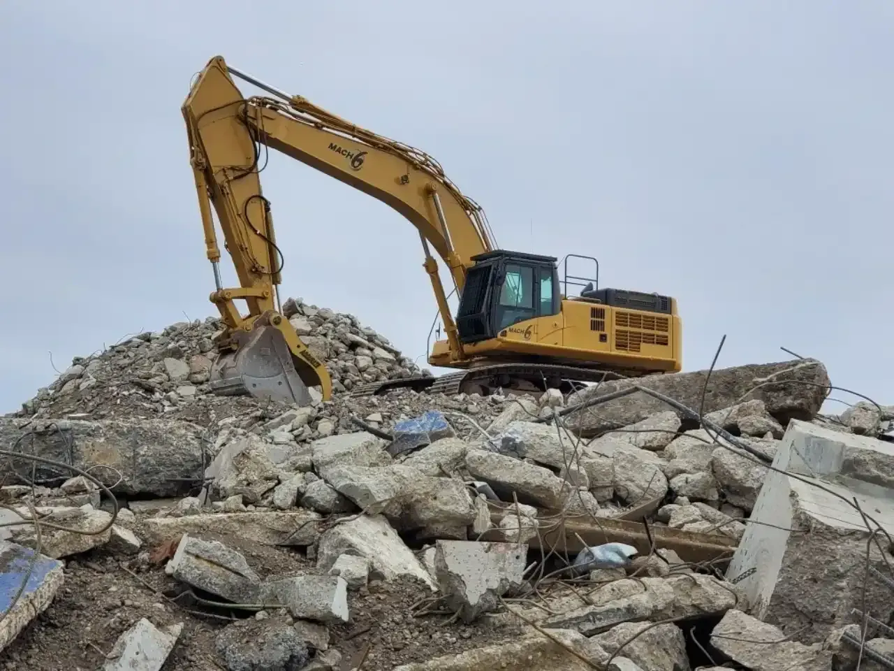 Yellow excavator on a pile of concrete debris, under a cloudy sky.
