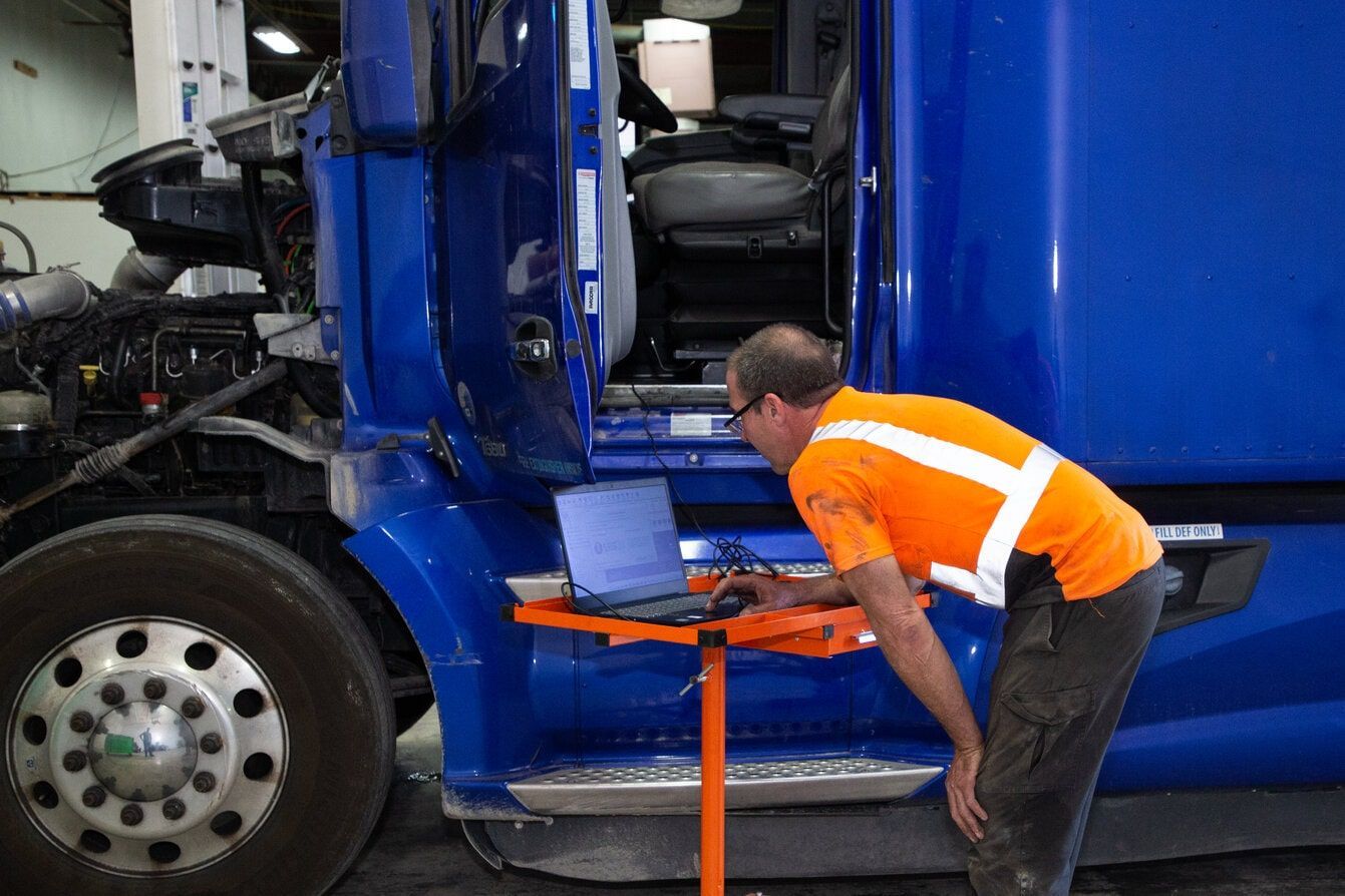 Mechanic using a laptop to diagnose a blue semi-truck in a garage, wearing safety vest.