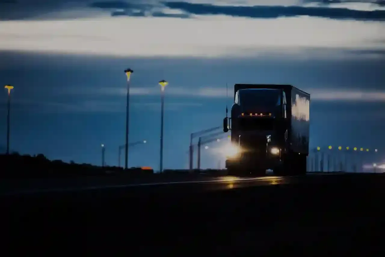 Semi-truck driving on a road at dusk; lit streetlights and a dark blue sky in the background.