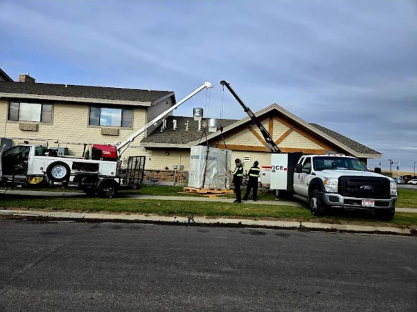 Cranes lifting a large rectangular unit beside a building with two workers watching on a grassy verge.
