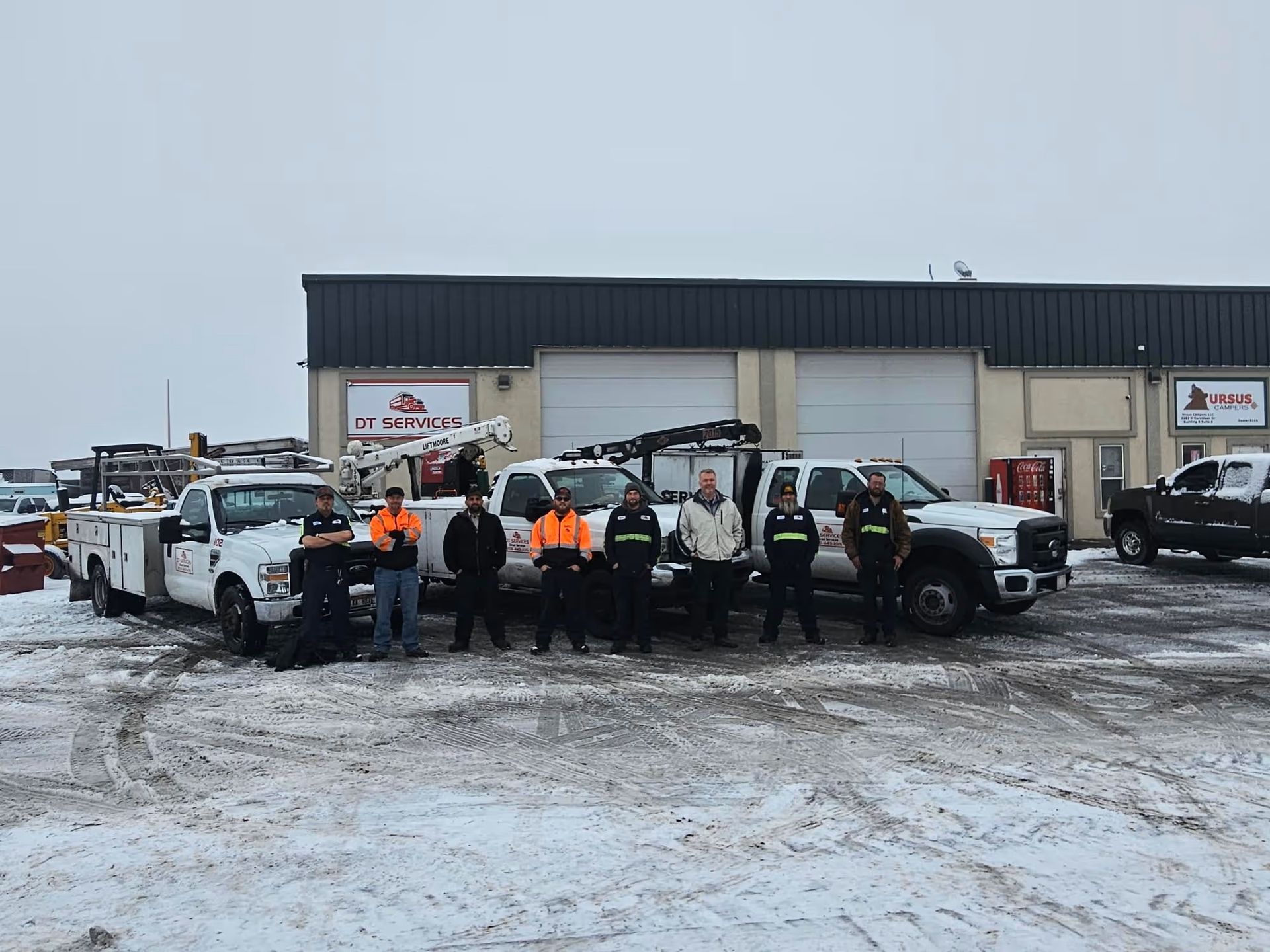 Group of people standing with tow trucks in snowy parking lot, in front of a building.
