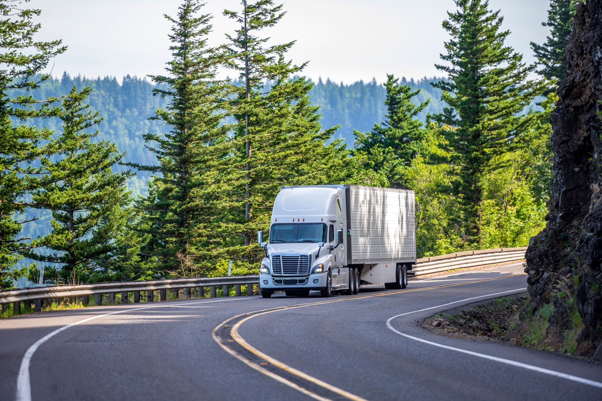 White semi-truck drives on a winding road through a forest.