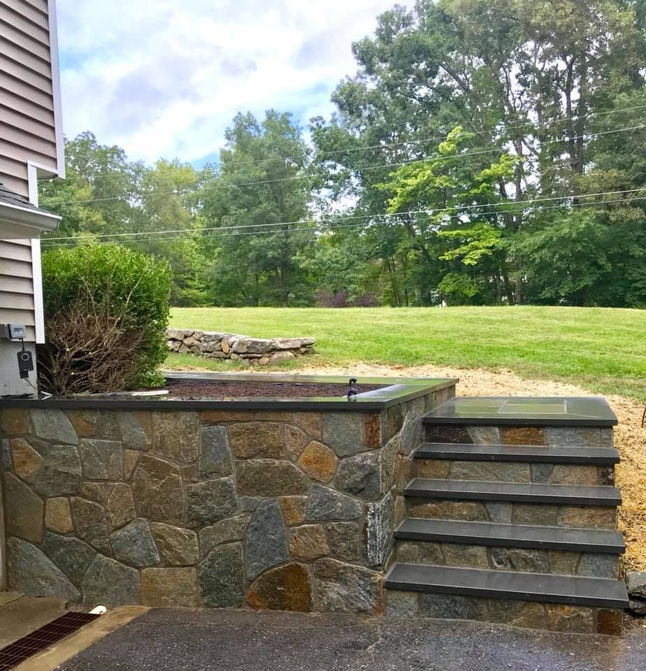 Stone wall with steps leading to a grassy yard; house on the left, trees in the background.