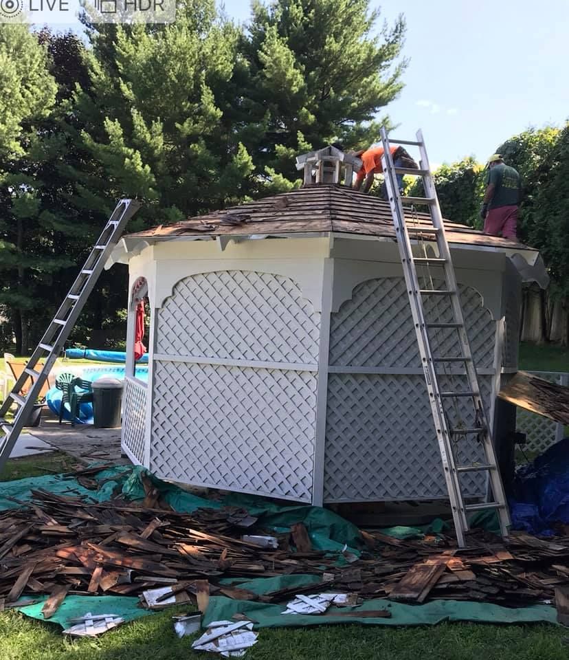 Men replace shingles on a white gazebo roof, surrounded by debris, ladders, and green foliage outdoors.
