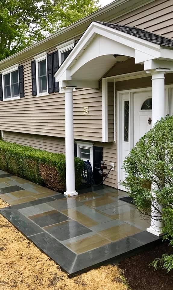 A front porch with bluestone walkway, columns, and a green hedge, beige siding.