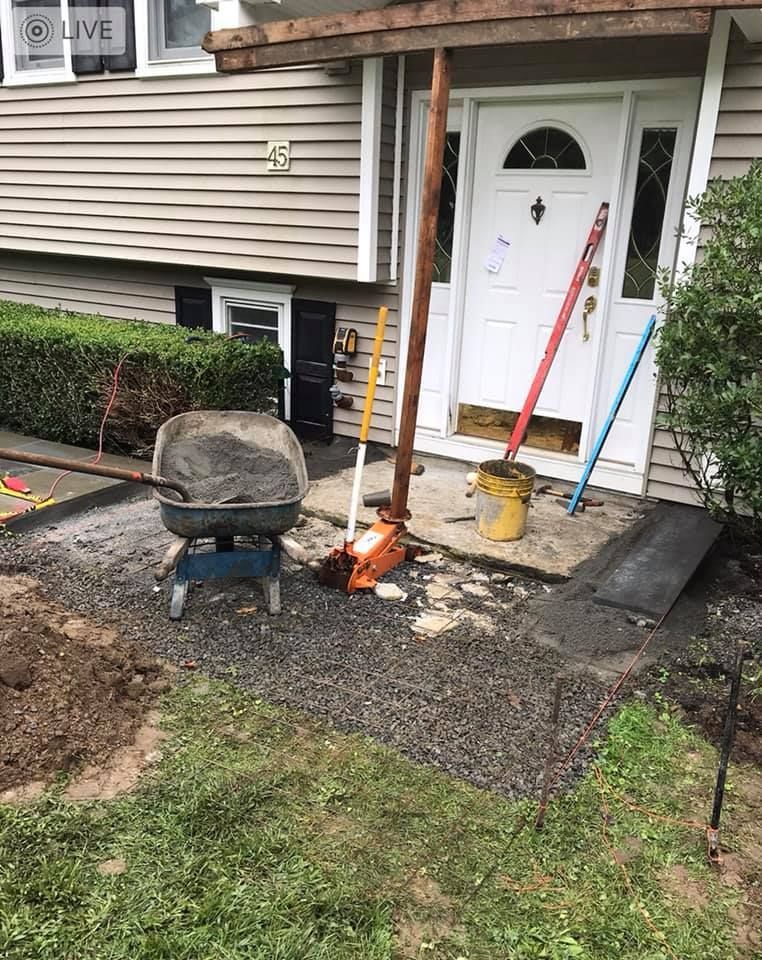 Construction site in front of a house: wheelbarrow, tools, gravel, unfinished walkway, support posts, and white door.