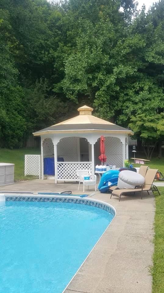 White gazebo by a pool, with blue water, a red umbrella, and green trees in the background.