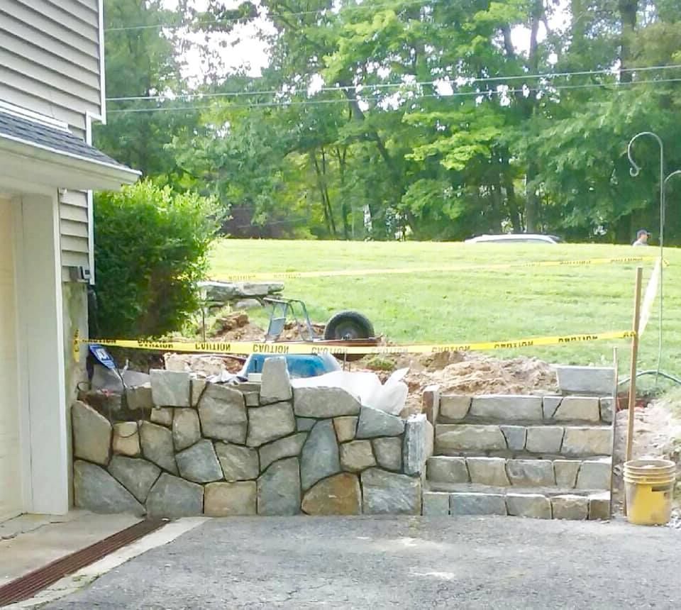 Stone retaining wall with steps under construction near a garage and grassy yard.