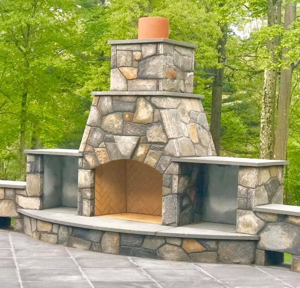 Outdoor stone fireplace with side counters, set on a stone patio, surrounded by greenery.