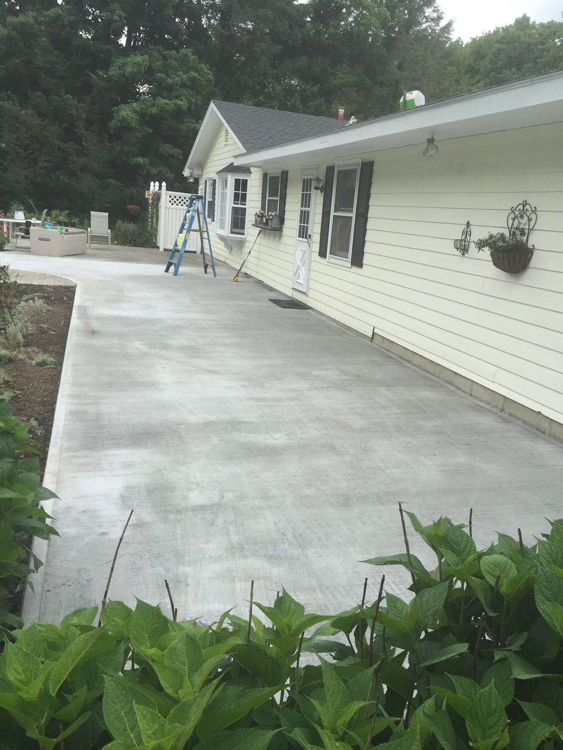 Light yellow house with concrete patio; blue ladder; greenery in front.