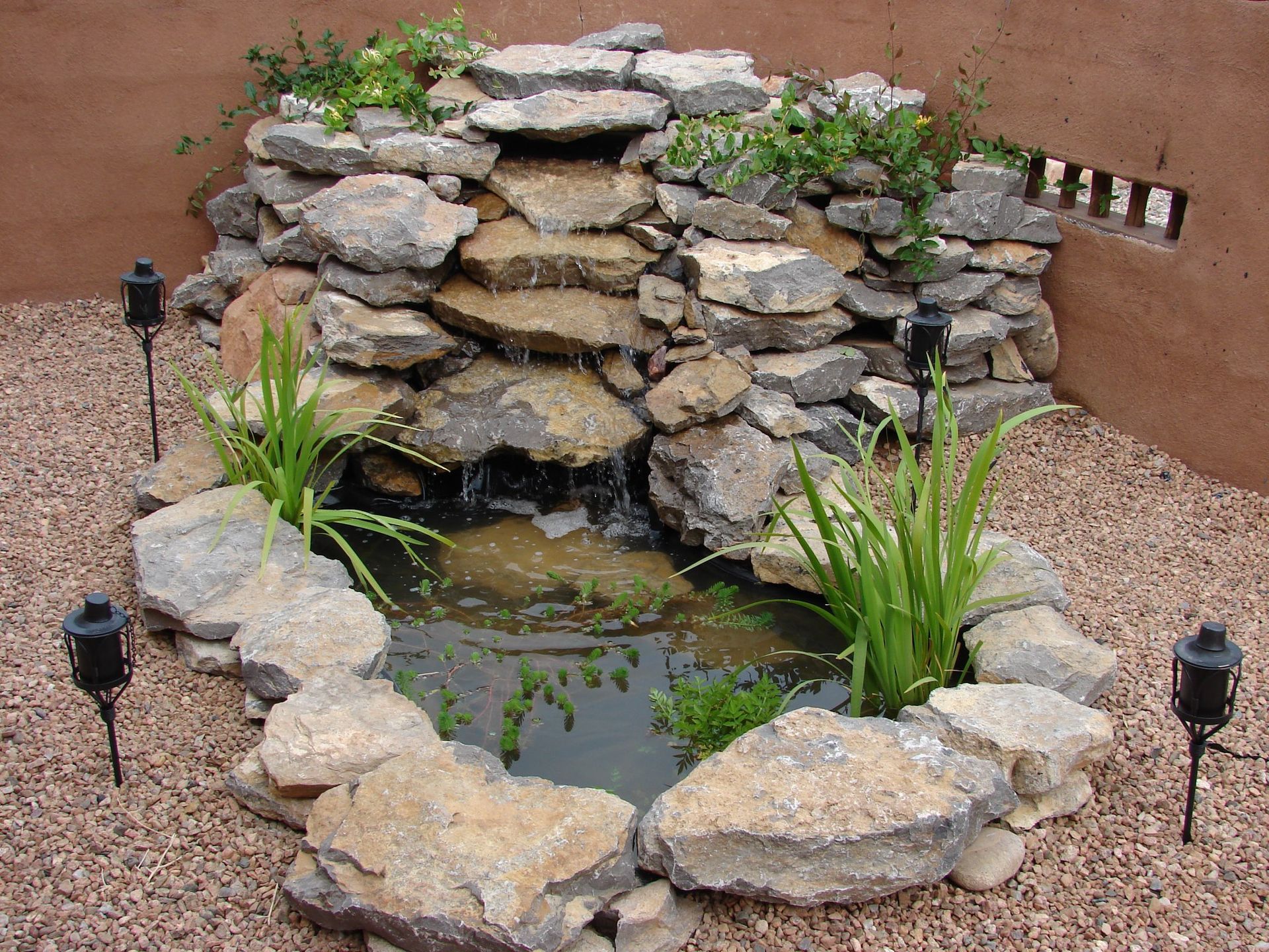 Small stone waterfall feature in a gravel-filled garden; water flows into a small pond, plants and torches surround it.