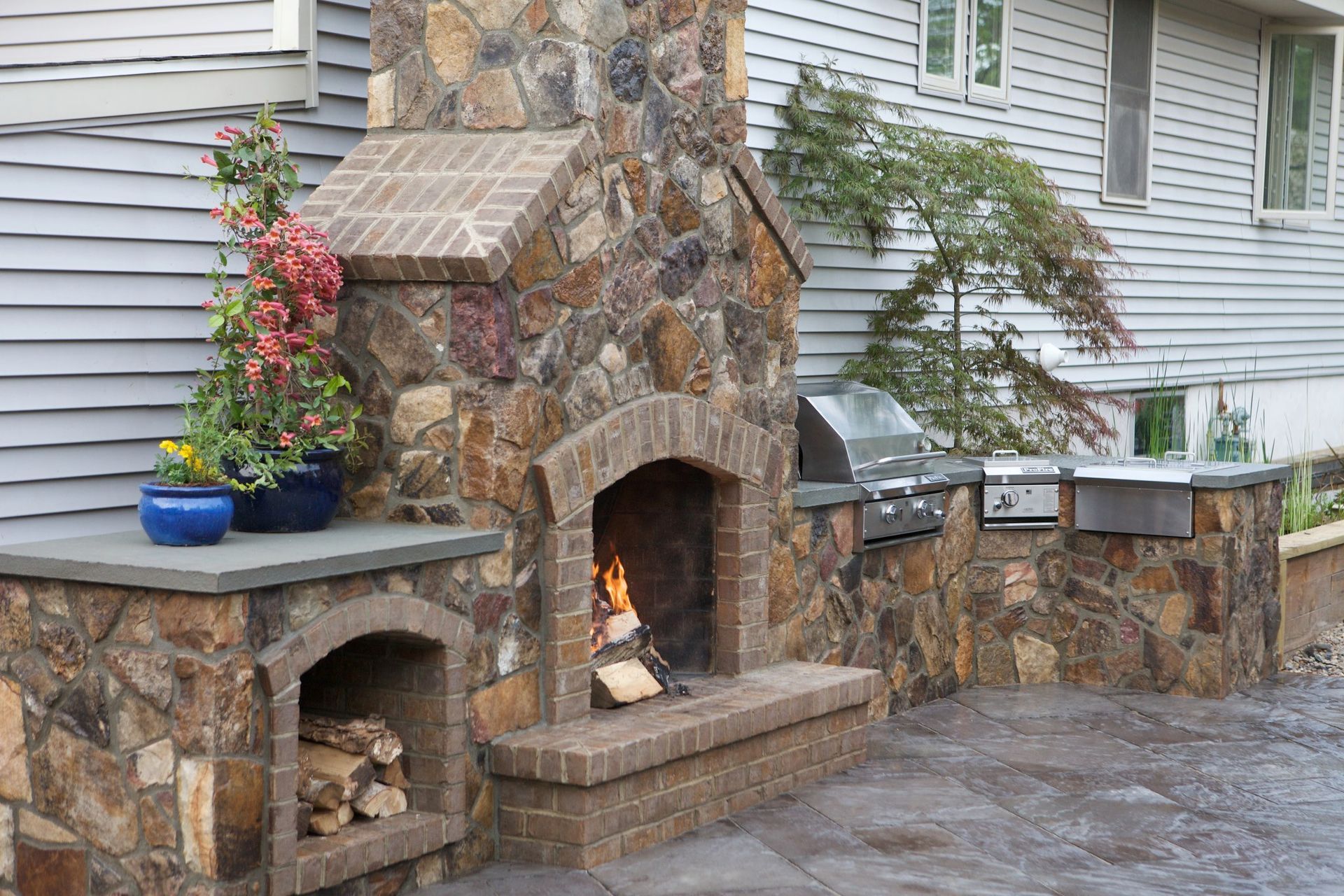 Outdoor stone fireplace with firewood storage, grill, and blue potted plants.