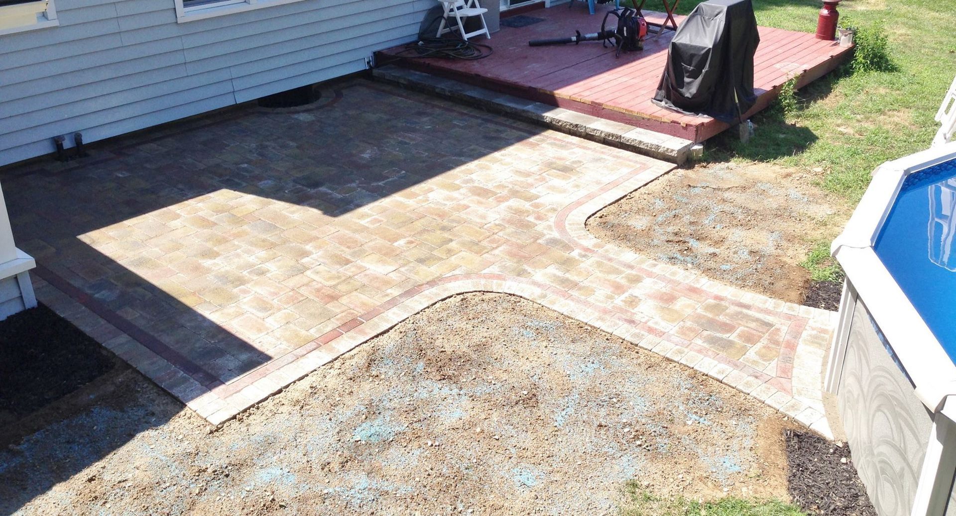 Brick patio and walkway next to a deck, pool, and house. Brown and tan tones.