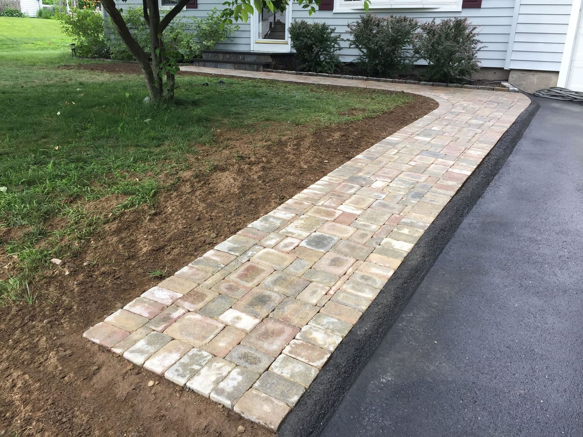 Brick walkway next to a driveway, leading to a house, surrounded by grass and landscaping.