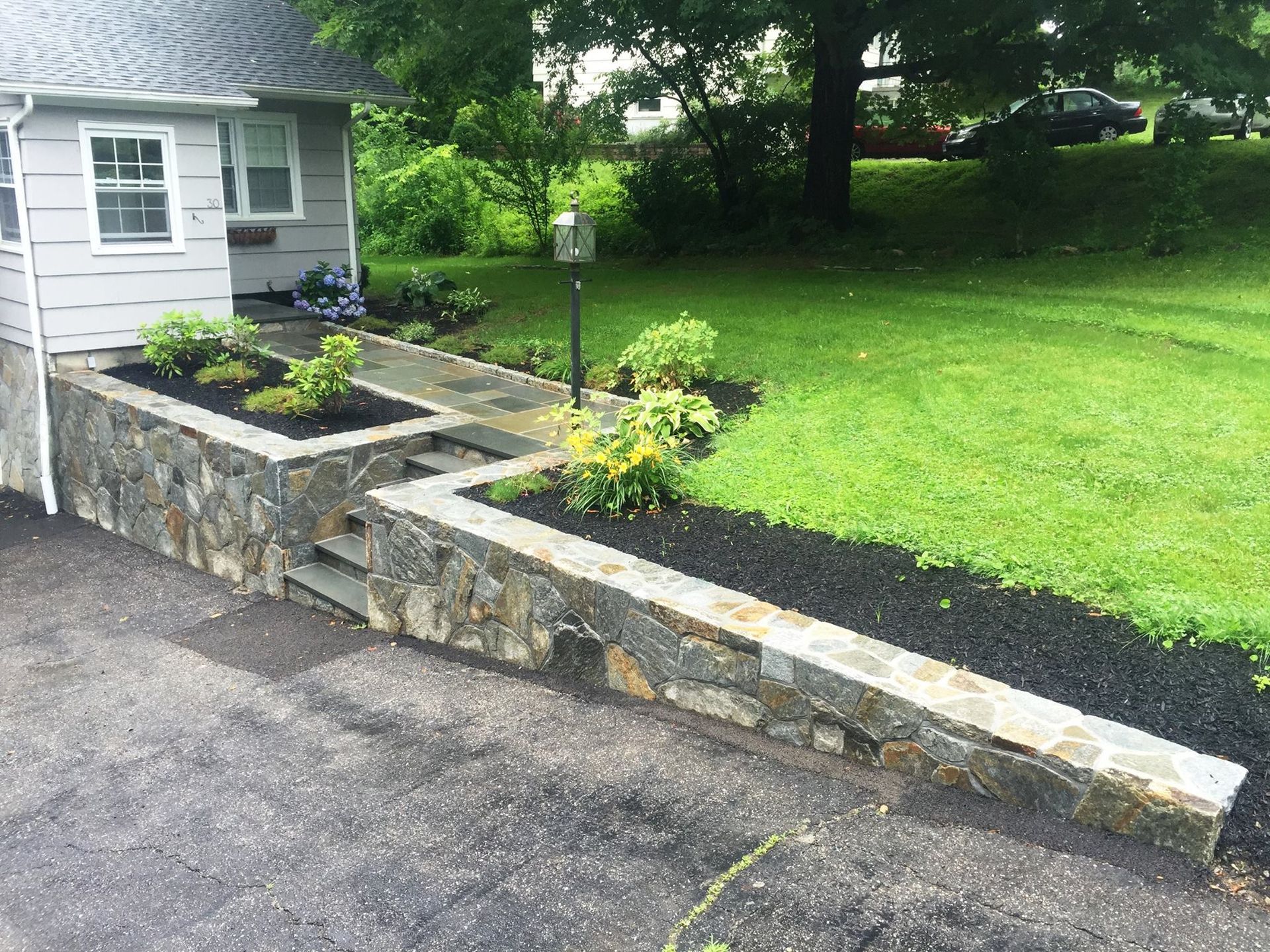 Stone retaining wall with steps and planters in front of a house with a lawn.
