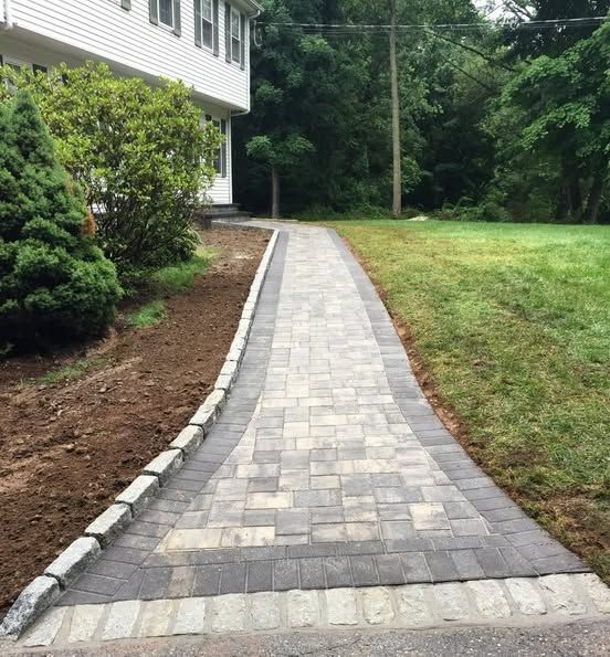 Brick walkway leading to a white house, bordered by a dark stone and grass.