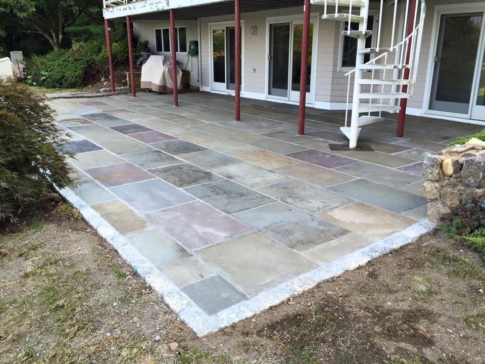Stone patio with various colored rectangular pavers, adjacent to a building with spiral staircase and grassy lawn.