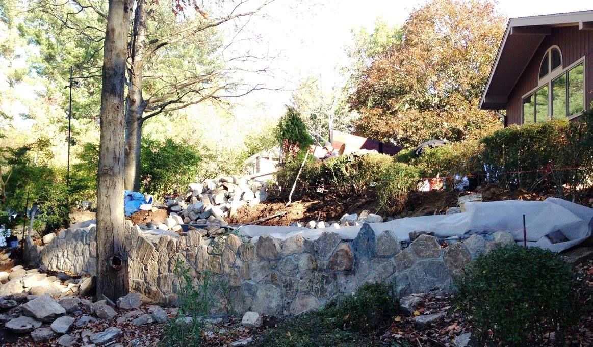 Stone wall bordering a yard with trees, shrubs, and a house in the background.
