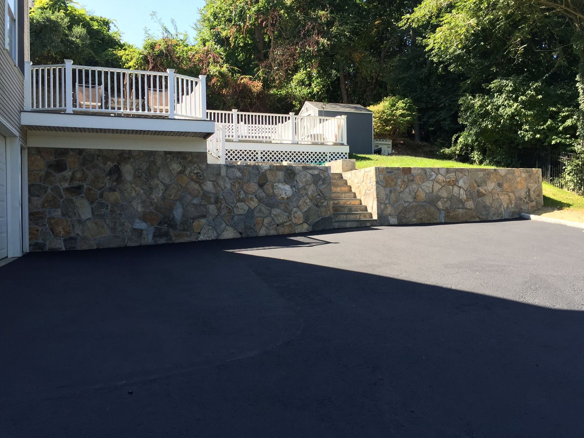 Asphalt driveway in front of a stone retaining wall with steps leading up to a deck.